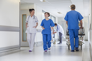 Photo of medical personnel walking through hallway, a woman in a white lab coat and a woman in blue scrubs are having a conversation while a man in blue scrubs pushes a patient bed down the hallway