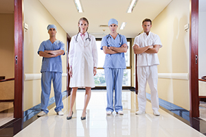 Photo of four hospital personel standing in a hallway from left to right a man in blue scrubs, a doctor with a lab coat, another man in scrubs with arms crossed and a man in white scrubs to the far right.