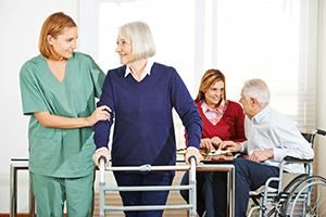 Photo of woman in scrubs helping elderly woman on a walker with two other patients in background talking to each other